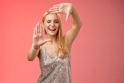 Sassy good-looking creative european blond woman in silver glittering dress winking cheeky smiling confident search inspiration around make frame hand search location take cool shot, red background.