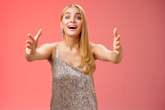 Excited charmign touched hearwarming young blond woman in silver party glamour dress stretch hands towards camera amused wanna hug cuddle hold cute puppy arms, standing red background.