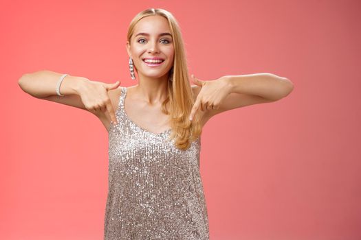 Lifestyle. Attractive glamour blond woman in silver glittering dress pointing down smiling excited showing awesome party place hang-out inviting try-out standing pleased red background enjoying perfect evening.