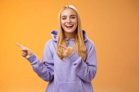 Charismatic firendly good-looking blond young female student pointing left discussing awesome new restaurant suggesting friend go check out laughing smiling happily, orange background.