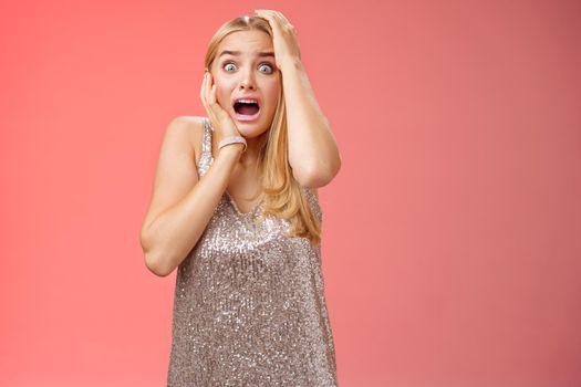 Shocked terrified blond woman horrified see crime screaming pop eyes shouting hold hands head afraid trembling fear standing frightened drop jaw gasping face terrible accident, red background.