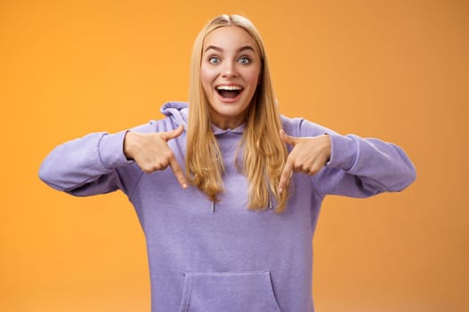 Excited fascinated european cute young girl blond hair smiling open mouth amused telling look down pointing downwards amazing cool new product recommend advertisement, orange background.