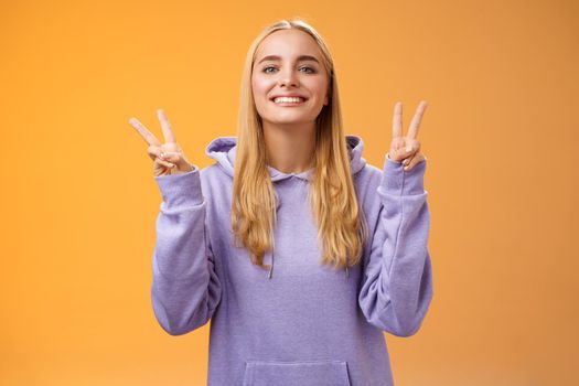 Lifestyle. Peaceful glad attractive blond european young female college student in hoodie smiling friendly make peace victory gesture spread love and happiness standing hippie sign orange background.