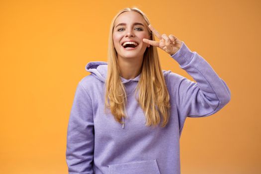 Attractive friendly carefree millennial blond girl in purple hoodie having fun friends laughing joyfully show peace victory hippie sign adore perfect chilly spring weather, orange background.