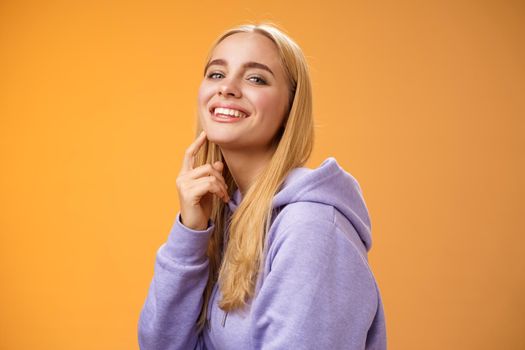 Attractive sassy self-assured caucasian blond female in purple hoodie turning camera touching chin cheeky smile camera check out interesting thing gazing forward intrigued, standing orane background.