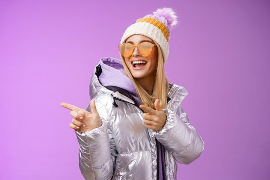 Hey how you doin. Cheeky stylish bond girl having fun greeting friend pointing finger pistols left smiling sassy say hello what up wearing cool silver jacket hat sunglasses, purple background.