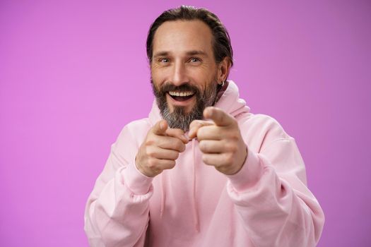 Funny amused carefree happy adult handsome man pranking friend fool around pointing camera index fingers greeting choosing you laughing joyfully having fu, standing purple background rejoicing.