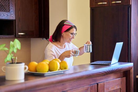 Woman in the kitchen with a cup of coffee looking into the laptop screen. Middle aged woman using video call for meeting and morning coffee. Technology, lifestyle, home, mature people concept