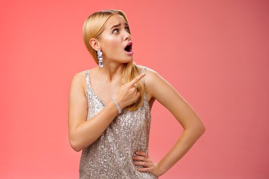 Shocked displeased bothered arrogant blond glamour woman in silver glittering dress turning upper right corner pointing complaining strange noise come upstairs, standing questioned red background.