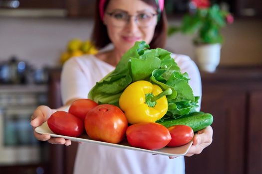 Close-up of a plate with tomatoes, cucumbers, paprika and lettuce in the hands of a woman. Kitchen interior background, fresh organic vegetables, home cooking, vegetarianism, nutrition, diet concept