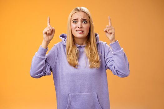 Worried perplexed unsure cute blond woman presenting project standing awkward afraid humiliation crining scared pointing up raised index finger waiting reply nervously, orange background.