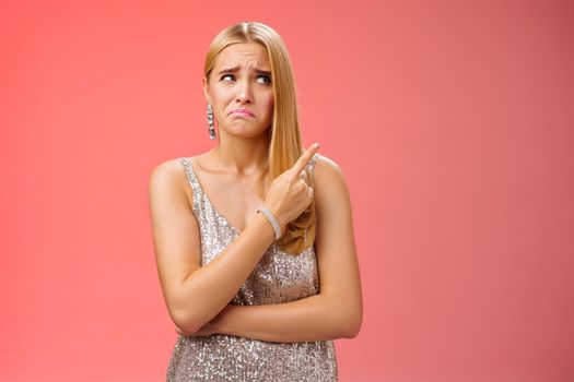 Upset doubtful unsure cute blond funny glamour girl in silver evening dress cringing grimacing hesitant pointing looking upper right corner suspicious, uncertain standing red background.