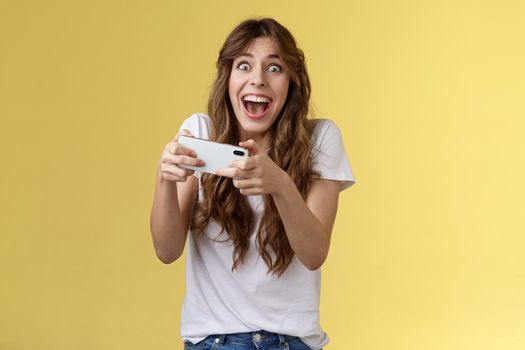 Extremely happy thrilled playful girl gamer playing awesome great new smartphone game hold mobile phone horizontal cheering look camera astonished impressed beating record yellow background.