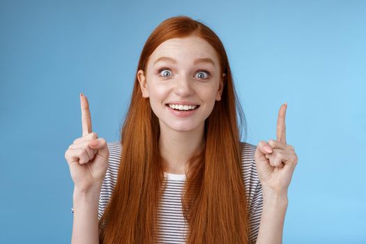 Thrilled happy young rejoicing redhead girl get awesome chance look surprised astonished smiling broadly wide eyes impressed incredible offer pointing up index fingers excitement, blue background.