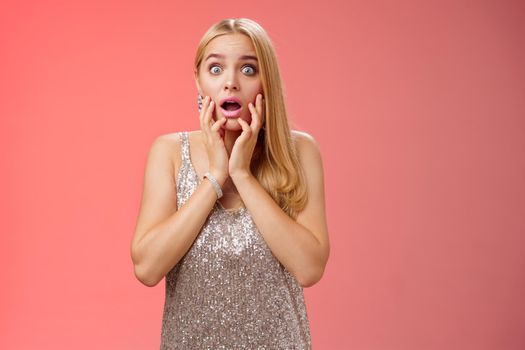 Shocked concerned stunned young european blond woman in silver dres drop jaw widen eyes stunned speechless gasping open mouth touch face amazed, look troubled perplexed, red background.