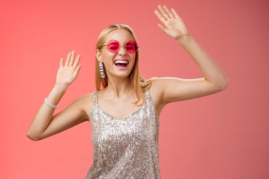 Happy amused carefree blond woman go wild dance-floor dancing having fun yelling yeah closed eyes waving hands moving rhythm music joyfully party in silver stylish dress sunglasses, red background.