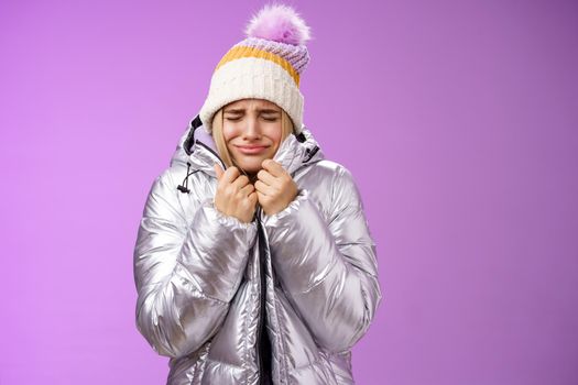 Unhappy sobbing whining cute blond girl pull jacket tight body close eyes crying freezing cold standing snowy winter resort shaking low temparature, purple background suffering discomfort.