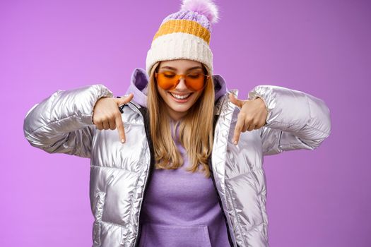 Joyful stylish blond woman in silver jacket stylish sunglasses jat enjoying perfect mountains view snowy vacation resort look pointing down smiling amused having fun feel happy, purple background.