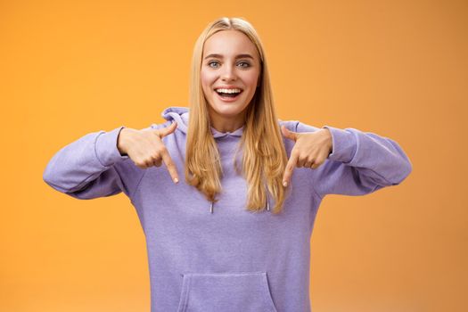 Amused joyful nice blond girlfriend pointing down present cool new product smiling broadly recommending try check out standing orange background happily grinning in hoodie. Advertisement concept