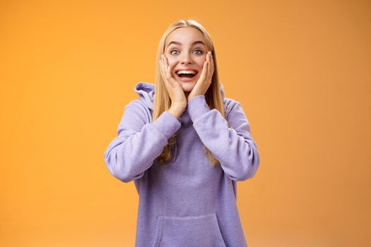 Surprised happy astonished fair-haired girlfriend receive incredible super cute gift touch cheeks amused reacting satisfied impressed standing excited orange background in casual hoodie.