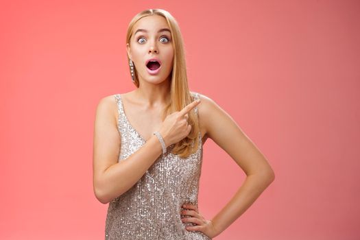 Lifestyle. Amused speechless impressed european blong girl drop jaw widen eyes thrilled pointing upper right corner excited amazed standing red background gossiping wealthy bartender wear silver shiny dress.