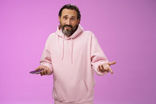 So what. Confused questioned adult bearded stylish guy in pink hoodie shrugging hands spread sideways dismay holding smartphone look clueless unaware camera not know how deal annoying phone calls.