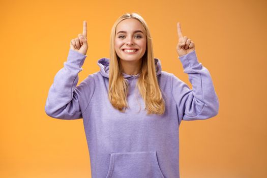 Friendly charismatic energized young blond european woman in cozy hoodie raise hands pointing up index fingers presenting awesome adverisement smiling happily recommend product, orange background.