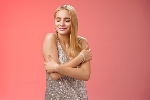 Lifestyle. Tender feminine elegant blond young woman in stylish silver glittering dress close eyes dreamy smiling touching herself cuddle embrace hands crossed body remember heartwarming passionate feelings.