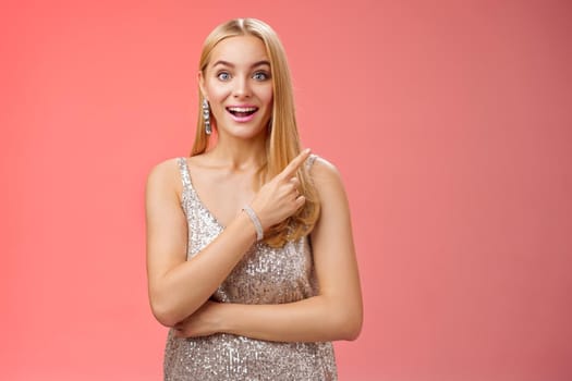 Joyful surprised good-looking blond woman talking friend during party smiling broadly look camera amused wondered pointing upper right corner curious discuss awesome room design, red background.