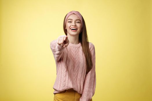 Amused charming tender young european girlfriend mocking having fun of someone pointing finger at camera laughing over funny situation, chuckling, mockering against yellow background. Copy space