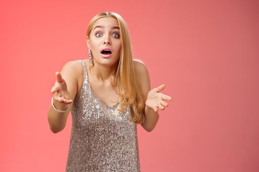 Impressed overwhelmed blond woman retelling exciting story gesturing astonished widen eyes shrugging explaining what happened talking passionately in silver glamour dress standing red background.