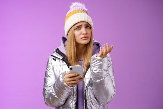 Questioned complicated cute blond girlfriend receive strange message look perplexed confused raising hand shrugging lift eyebrow cannot understand meaning holding smartphone, purple background.