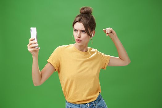 Woman making serious look to take selfie while listening music in wireless earphones posing and staring at smartphone screen daring and aggressive, liking post photos online over green background. Technology and lifestyle concept