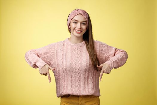 Tender young 20s european girl in sweater smiling broadly finally take-off braces pointing down promoting skillful dentist professional. Female student standing happy positive over yellow wall.