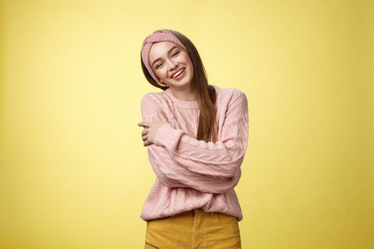 Cozy charming friendly young smiling girl in knitted warm comfortable sweater, grinning joyfully tilt head emracing herself, crossing arms, hugging feeling safe and happy, posing against yellow wall.