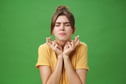 Worried intense, concerned attractive young european female model in yellow t-shirt raising shoulders crossing fingers for good luck pursing lips and closing eyes while waiting for miracle hopefully. Body language concept