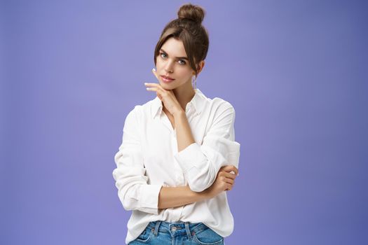 Portrait of tender and feminine stylish woman in white blouse posing sensually and flirty touching chin gazing daring at camera posing against purple background with self-assured expression. Copy space