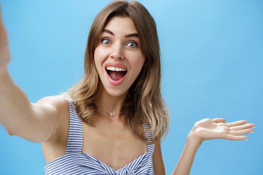 Waist-up shot of charismatic upbeat enthusiastic cute female blogger raising hand over copy space and holding camera as if recording video from vacation showing cool product over blue background. Lifestyle.