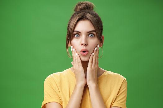 Portrait of impressed and shocked cute feminine woman in yellow t-shirt folding lips from excitement and interest touching cheeks surprised reacting to amazing rumor posing over green background. Body language concept