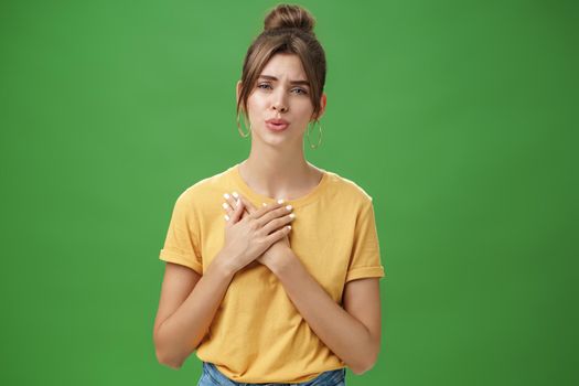 Touched and pleased attractive young woman in yellow t-shirt holding palms pressed to chest in thankful and grateful gesture folding lips melting heart seeing cute thing over green background. Copy space