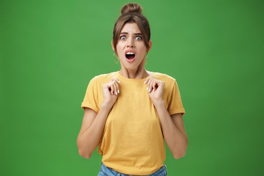 Shocked feminine glamour woman with combed hair in yellow t-shirt frowning gasping with opened mouth holding palms pressed to chest from aversion and disgust looking at creepy thing over green wall. Body language concept