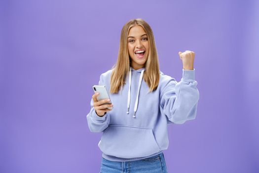 Yes we did it. Pleased excited woman in hoodie raising fist in celebration and success gesture smiling broadly triumphing holding smartphone reacting to positive news in cellphone over purple wall. Technology and emotions concept