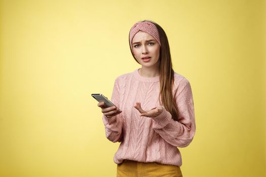 Puzzled confused upset questioned young cute girl in headband knitted sweater frowning clueless raising hand in dismay holding smartphone, cannot understand what happened cellphone over yellow wall.