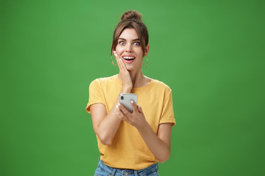 Surprised touched and impressed beautiful european girl with diastema smiling broadly touching cheek from amazement and joy holding smartphone looking happy at camera after reading cool news. Technology concept