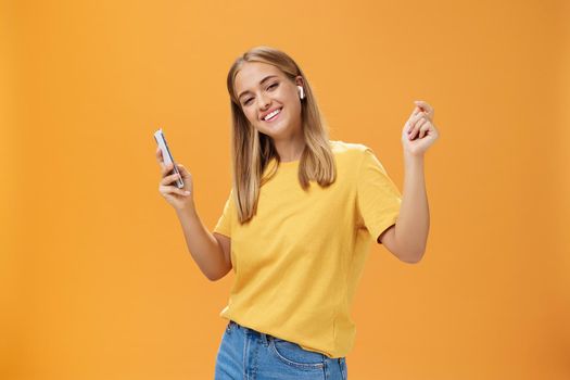 Joyful carefree european woman wearing wireless earbuds holding smartphone dancing with raised hands and pleased happy smile leading vanlife having time to relax and enjoy life over orange wall. Technology concept