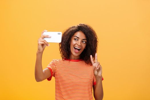 Smile to camera. Attractive and stylish self-assured dark-skinned female model with curly hairstyle showing peace gesture while taking selfie holding smartphone near face and smiling at gadget screen.