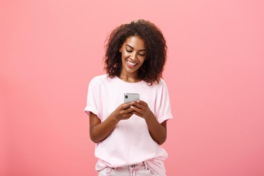 Girl writing text message to friend. Portrait of charming amused and happy young dark-skinned woman with curly hair looking at smartphone screen typing smiling enjoying cool features of cellphone.