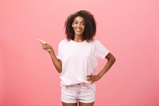 Friendly chill and relaxed attractive dark-skinned girlfriend with curly hairstyle holding hand on hip and pointing left with assured pleased expression showing where she bought cool outfit. Advertisement concept