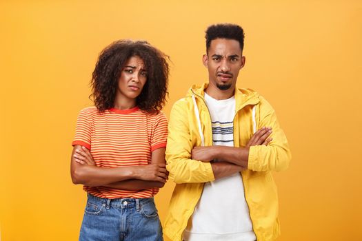 Two friends dislike lame idea of mate. Portrait of dissatisfied unimpressed african american man and woman crossing arms on chest in aversion frowning doubtful and disappointed over orange wall.