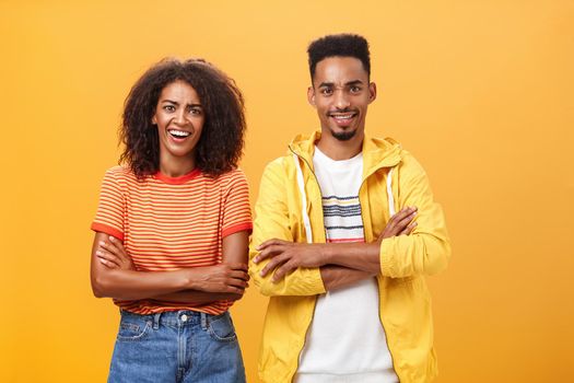 Portrait of surprised sarcastic african american woman with afro hairstyle standing with cute boyfriend crossing arms on chest laughing from scorn and fun over orange background. Copy space